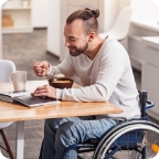 Man sits in wheelchair, eats breakfast and uses laptop.